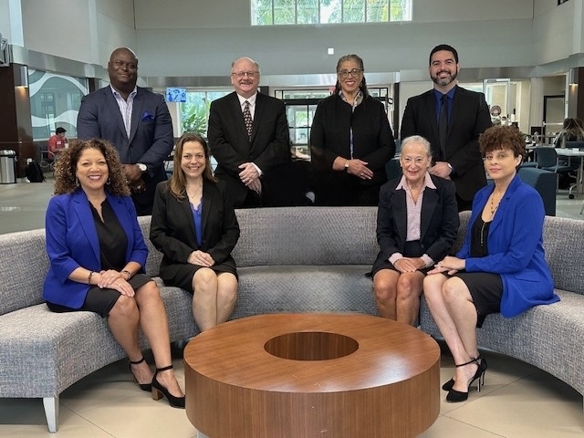 Group picture of smiling adults sitting on a gray couch and posing behind the couch